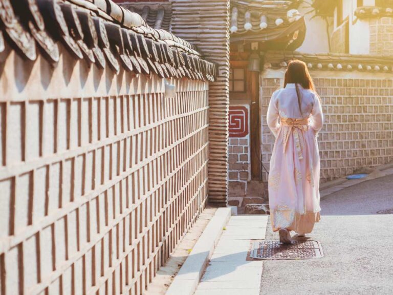 韓国・ソウルの北村韓屋村を、伝統衣装の韓服（ハンボク）を着て静かに歩く女性。柔らかな夕日が石壁と韓屋を温かく照らす、穏やかな時間の流れを感じる一枚。