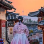 韓国・ソウルの北村韓屋村で、韓服を着た女性が夕焼け空の中を歩く風景。伝統家屋の街並みと遠くに見えるNソウルタワーが、ノスタルジックな雰囲気を演出している。