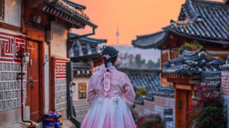 韓国・ソウルの北村韓屋村で、韓服を着た女性が夕焼け空の中を歩く風景。伝統家屋の街並みと遠くに見えるNソウルタワーが、ノスタルジックな雰囲気を演出している。