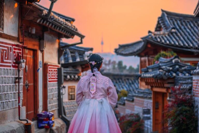 韓国・ソウルの北村韓屋村で、韓服を着た女性が夕焼け空の中を歩く風景。伝統家屋の街並みと遠くに見えるNソウルタワーが、ノスタルジックな雰囲気を演出している。