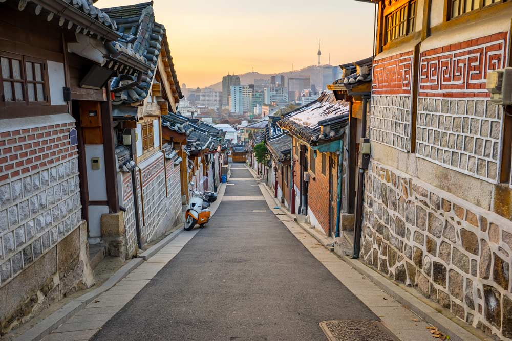 夕暮れ時の韓国・ソウル北村韓屋村の路地。伝統的な韓屋が並ぶ静かな通りの奥に、南山ソウルタワーとビル群が見え、ノスタルジックな雰囲気が漂う。