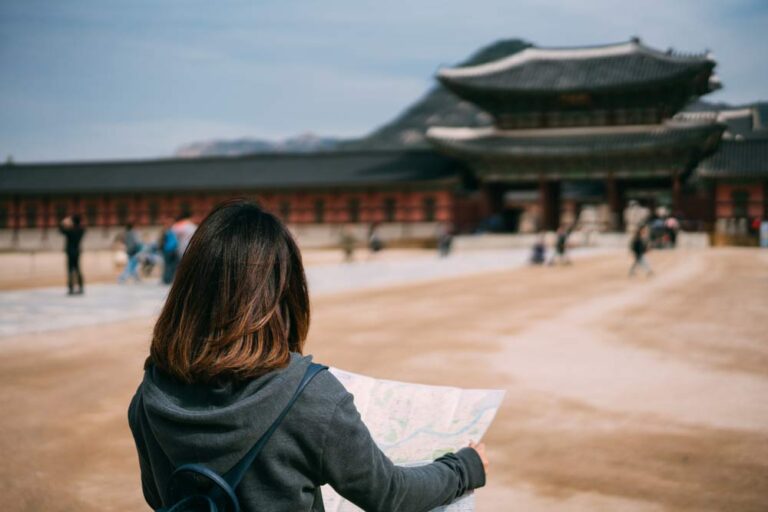 韓国・ソウルの景福宮を訪れ、地図を手に観光する旅行者の後ろ姿。伝統的な宮殿建築と背景の山々が調和する歴史ある風景。