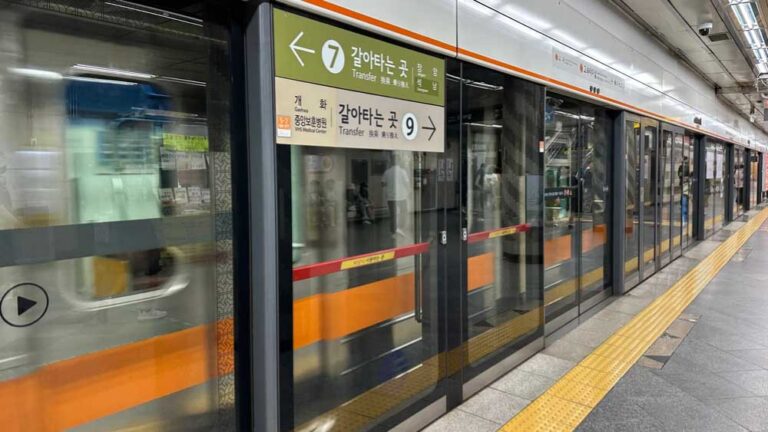韓国・ソウルの地下鉄5号線の駅ホーム。ガラスのホームドア越しに、乗客と到着する列車が見え、7号線と9号線への乗り換え案内表示も確認できる都市交通の一場面。