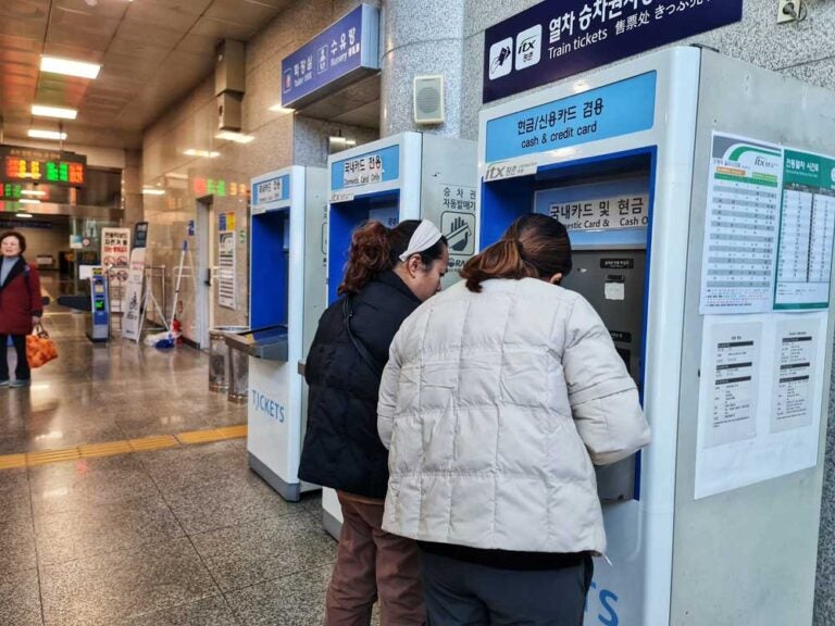 韓国の鉄道駅で自動券売機を利用する2人の女性。現金やクレジットカードに対応したiTXの切符販売機が並び、後方には時刻表表示や改札ゲートが見える。