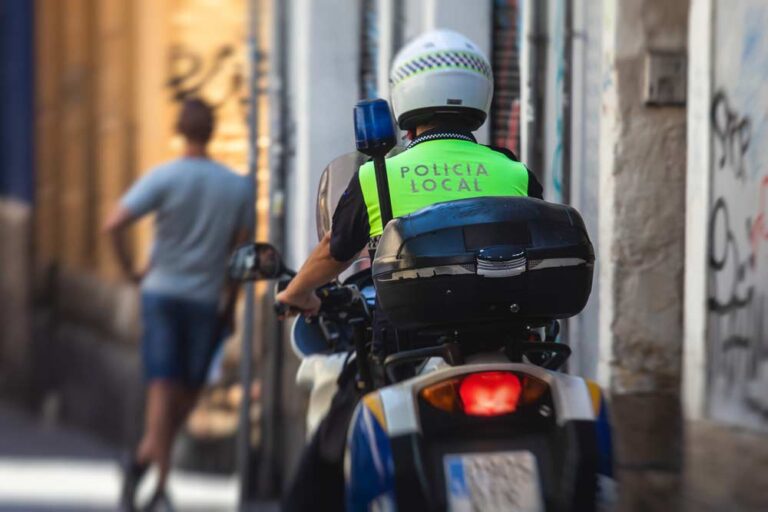 スペインの地方警察官がバイクで路地をパトロールしている様子。背中の「Policía Local」の文字が目立つ。