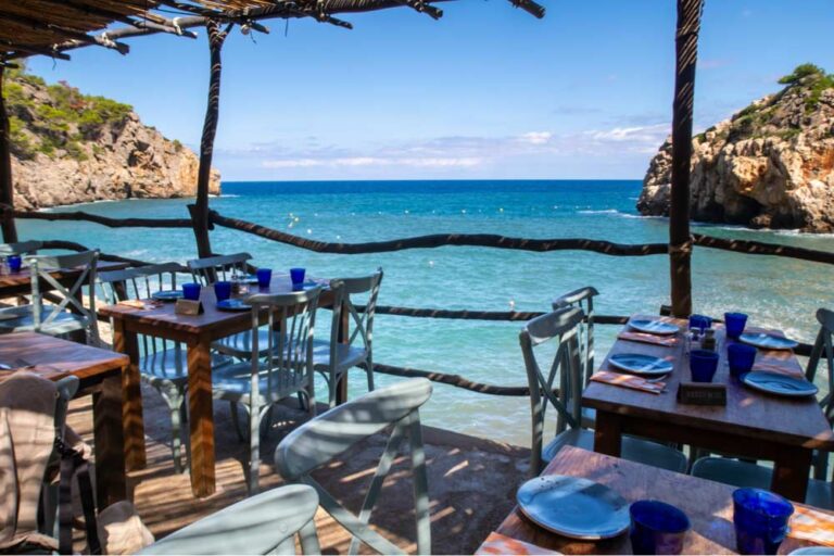 スペイン・マヨルカ島の入江沿いにあるオープンエアのレストラン。波の音をBGMに、青いグラスと木製テーブルが並ぶ、絶景を望む特等席のダイニングエリア。