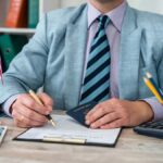 アメリカのビザ申請書に記入する男性。デスク上にはパスポート、アメリカ国旗、時計、スマートフォン、文房具が並び、申請手続きを行う様子が表現されている。