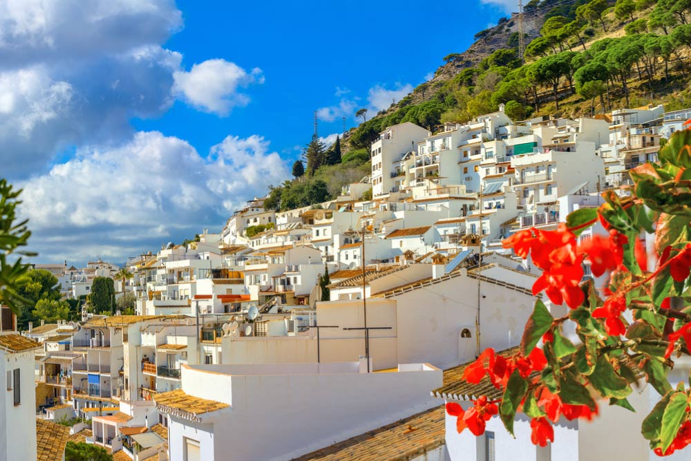 スペイン・ミハスの丘に広がる白い家々の街並み。青空と山の緑、赤い花が彩りを添える、アンダルシア地方の美しい白い村の風景。