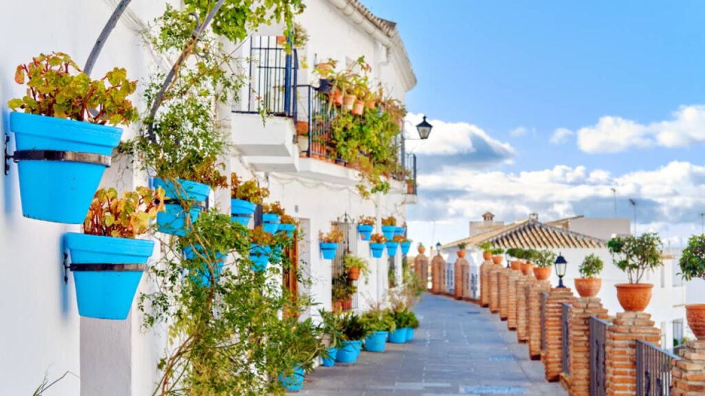 スペイン・ミハスの白い街並みに並ぶ鮮やかな青い鉢植え。白壁の家と色とりどりの植物、レンガの柵が美しく調和した、アンダルシアの伝統的な風景。