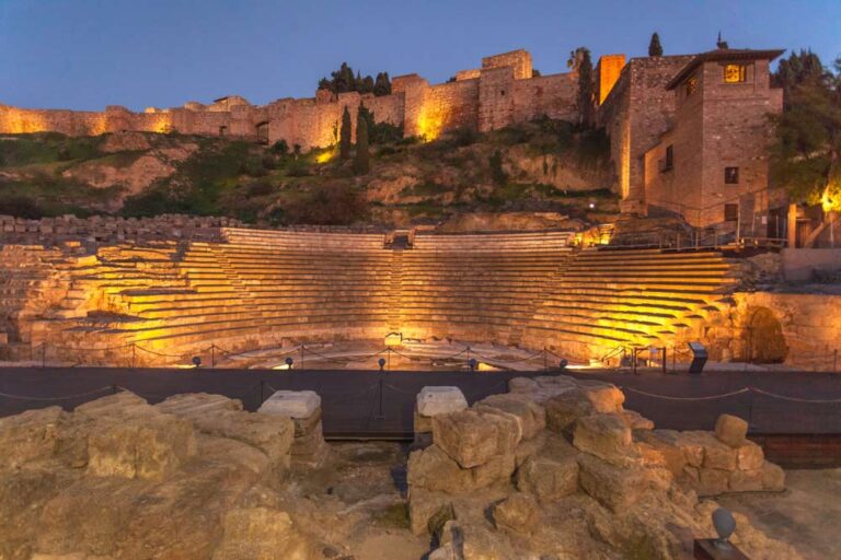 スペイン・マラガのローマ劇場（Teatro Romano de Málaga）を夕暮れ時にライトアップした風景。古代ローマ時代の円形劇場と背後に広がるアルカサバの城壁が幻想的に輝く歴史遺産。