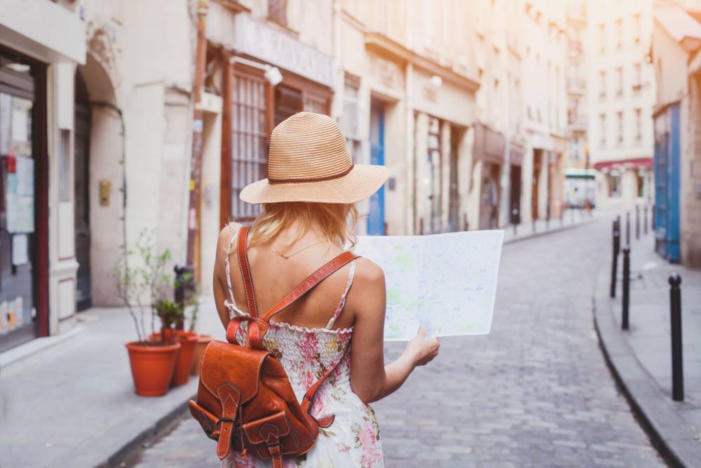 スペインの石畳の街並みを歩きながら地図を見る女性旅行者。麦わら帽子とレザーバッグを身に着け、異国情緒あふれる路地を散策する後ろ姿。