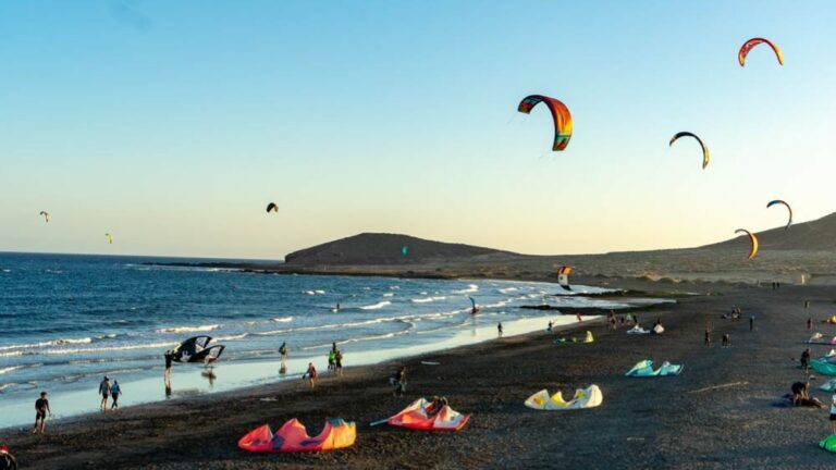 スペイン・テネリフェ島エル・メダノのビーチ。夕暮れ時、カイトサーフィンを楽しむ人々と、空を舞う色とりどりのカイトが広がる活気ある風景。