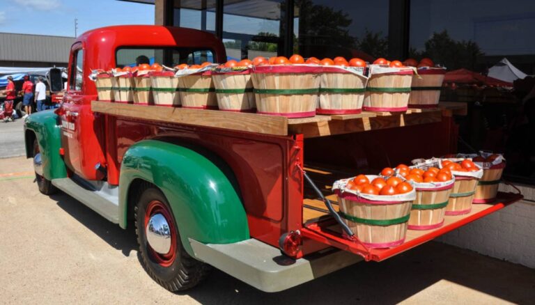 スペインの市場に停まるヴィンテージトラックの荷台に積まれたバスケット入りの新鮮なトマト。赤と緑のクラシックなトラックが農産物の彩りを引き立てる印象的な風景。