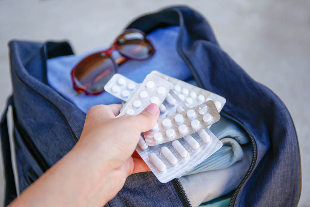 旅行用バッグに常備薬を準備する様子。サングラスや衣類と一緒に、錠剤パックを手に持っているシーン。