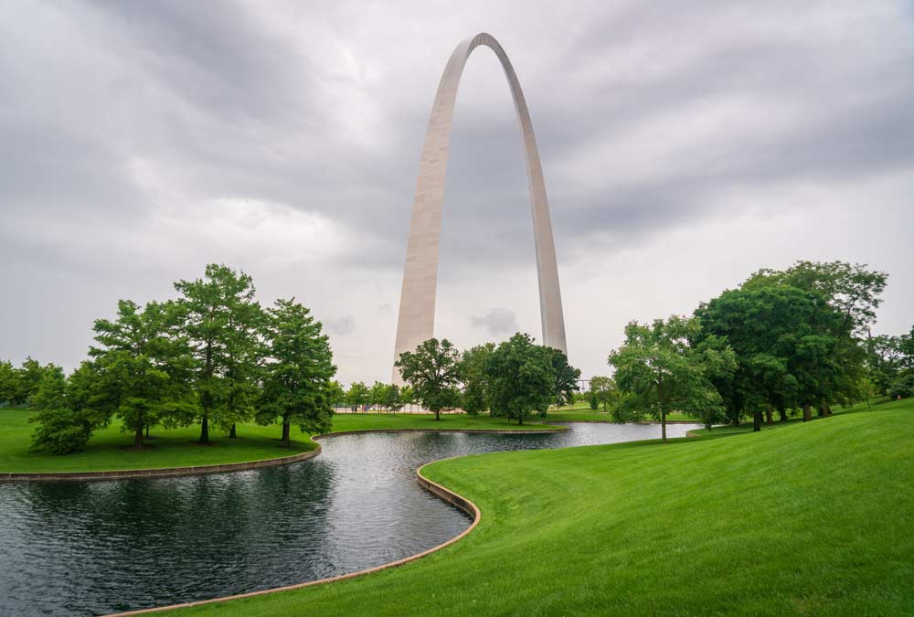 アメリカ・ミズーリ州セントルイスにあるゲートウェイ・アーチ。曇り空の下、緑豊かな公園と池に囲まれた美しい風景の中にそびえ立つ巨大なアーチ型モニュメント。