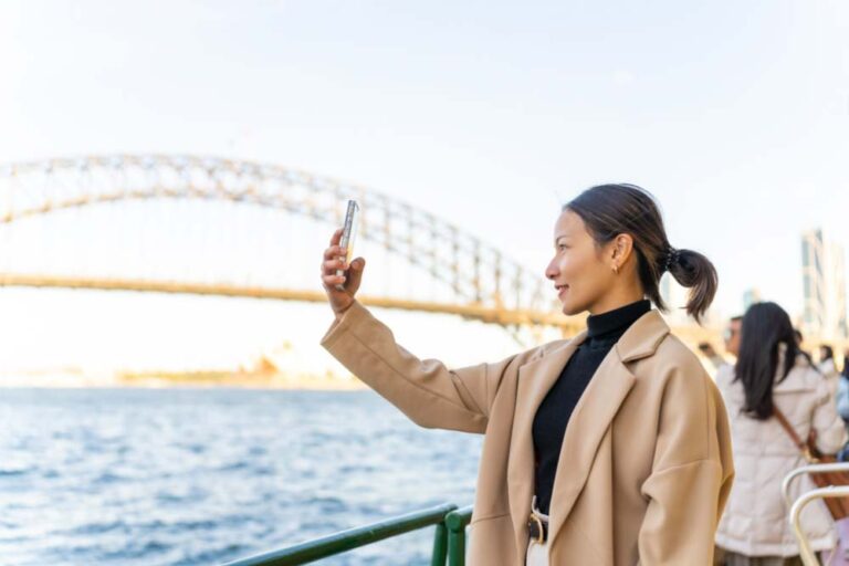 シドニーハーバーブリッジを背景に、フェリーの上でスマホを手に自撮りする女性旅行者。