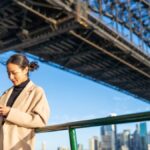 シドニーハーバーブリッジの下でスマートフォンを操作する女性。背景には青空とシドニー市街地が広がる。