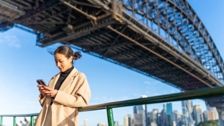 シドニーハーバーブリッジの下でスマートフォンを操作する女性。背景には青空とシドニー市街地が広がる。