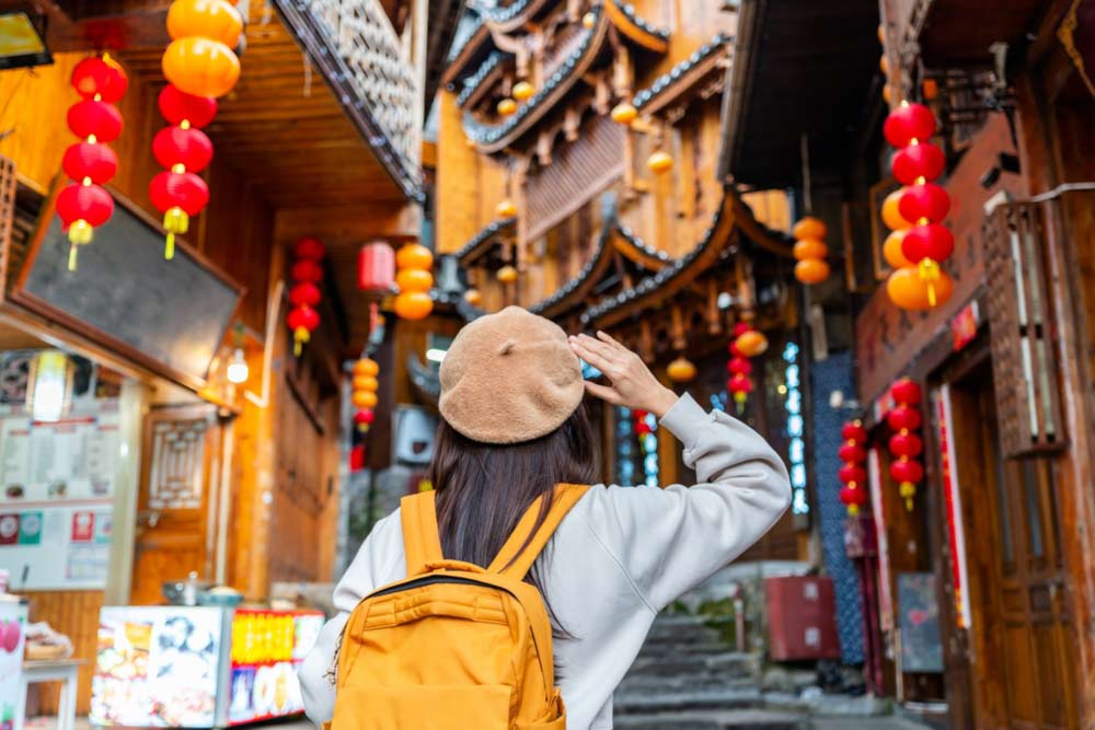 中国の伝統的な街並みを散策する黄色いリュックを背負った旅行者の後ろ姿。赤い提灯が並ぶ趣ある風景。