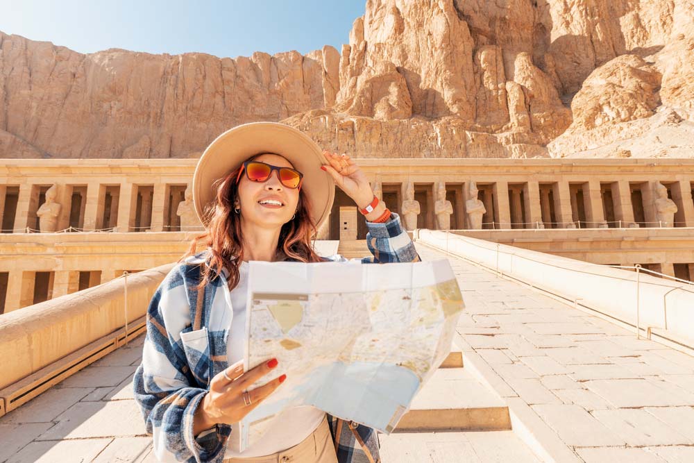 エジプト・ルクソールのハトシェプスト女王葬祭殿を背景に、地図を手に観光を楽しむ帽子とサングラス姿の女性旅行者。