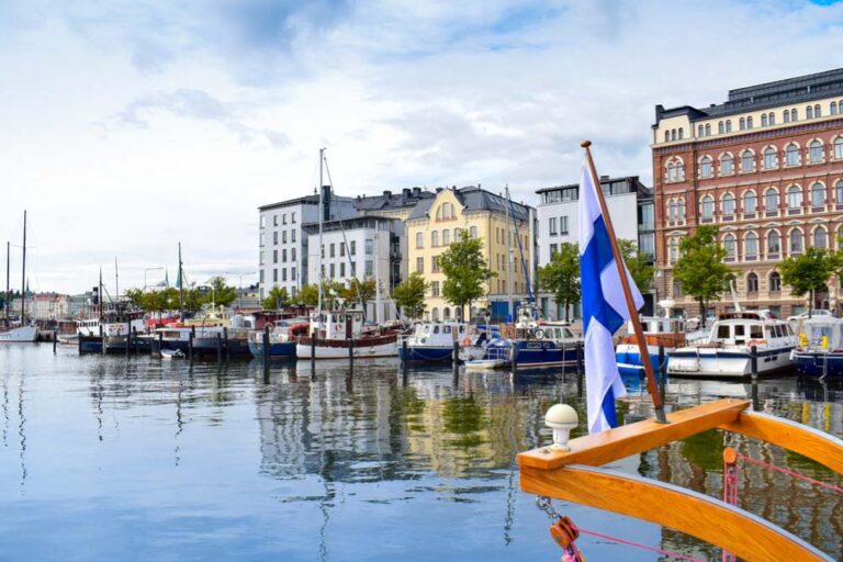 フィンランドの国旗が掲げられたボートと、ヘルシンキの港に停泊するヨットやカラフルな建物が並ぶ風景。