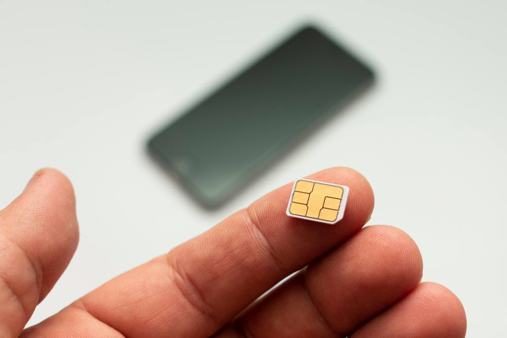 SIMカードを指で持つ手元のクローズアップ。背景にはぼやけたスマートフォンが置かれている。