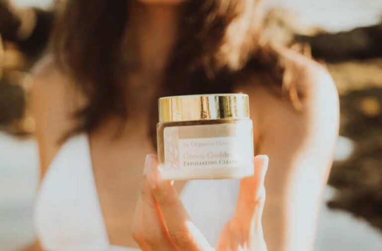 白いビキニを着た女性が、Ao Organics Hawaii の「Green Goddess Exfoliating Cleanser(グリーンゴッデス・エクスフォリエイティング・クレンザー)」の瓶を手に持っている。