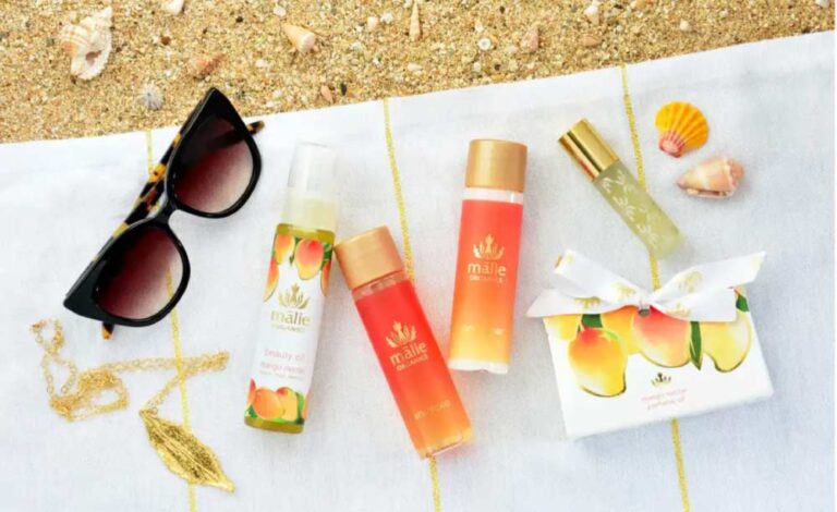 砂浜に敷かれた白いタオルの上に、マリエオーガニクスのマンゴーネクターシリーズのコスメ(ビューティーオイル、シャワージェル、トナー、パフュームオイル)、サングラス、貝殻、ゴールドのネックレスが並べられている。