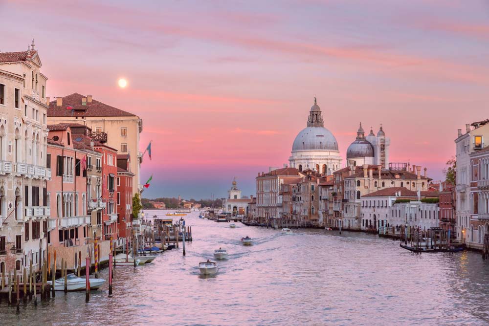 イタリア・ヴェネツィアのグランドカナル沿いに並ぶ歴史的建物と夕焼けに染まる空、運河を行き交うボートの風景