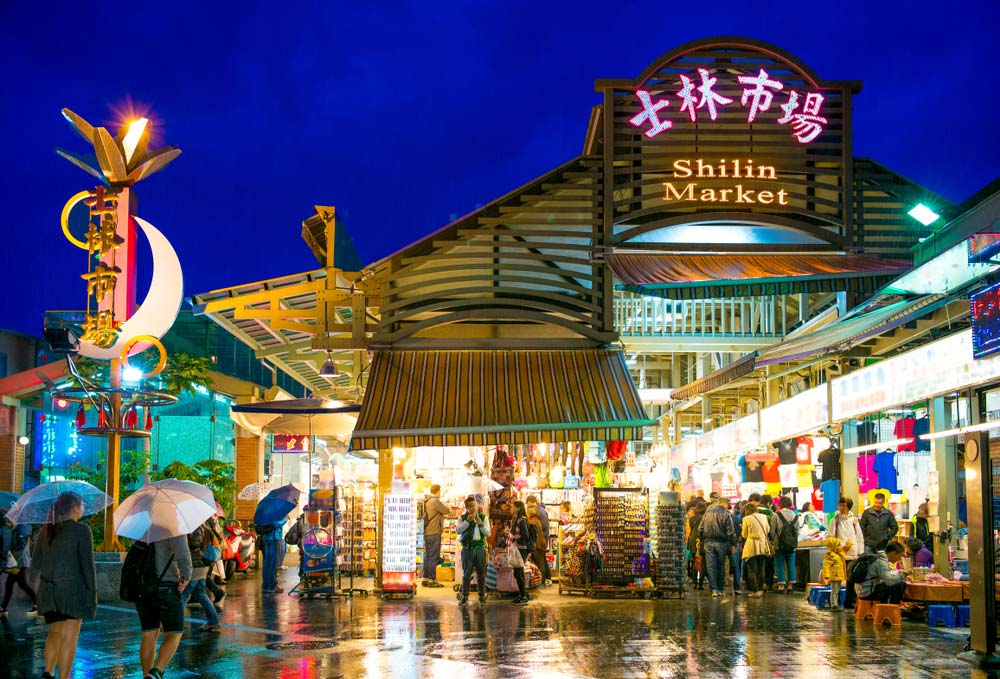 台北の士林夜市の入り口。雨の中でも多くの人で賑わい、傘を差しながらショッピングを楽しむ観光客の様子。