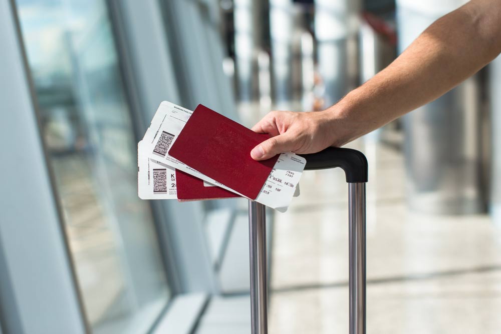 空港でスーツケースの取っ手に手をかけ、パスポートと搭乗券を持つ旅行者の手元。中国の観光ビザ手続きや渡航時のトラブルを想起させる場面。