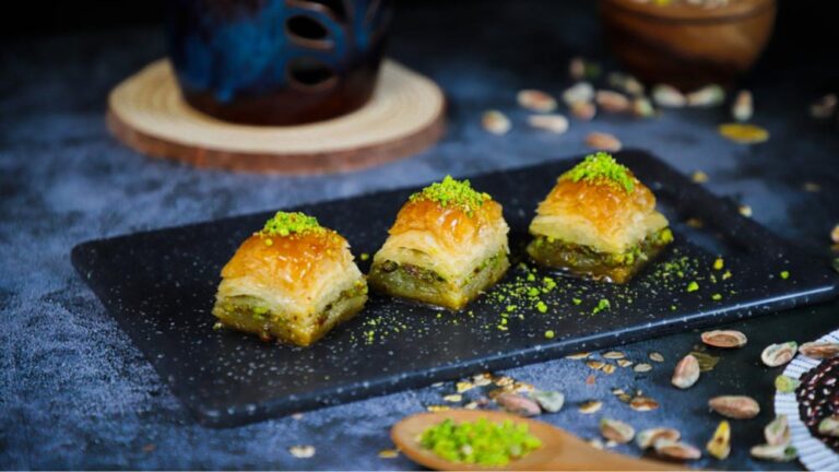 サクサクのパイ生地とピスタチオを層に重ねたトルコ伝統菓子バクラヴァが黒いプレートに3個並び、まわりにはピスタチオの粉とナッツが散りばめられている。