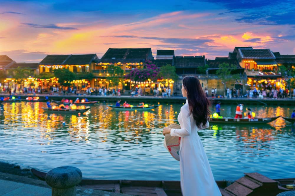 ベトナム・ダナン近郊のホイアンで、川沿いのランタンが灯る夕暮れの街並みを眺めるアオザイを身に纏った女性の後ろ姿