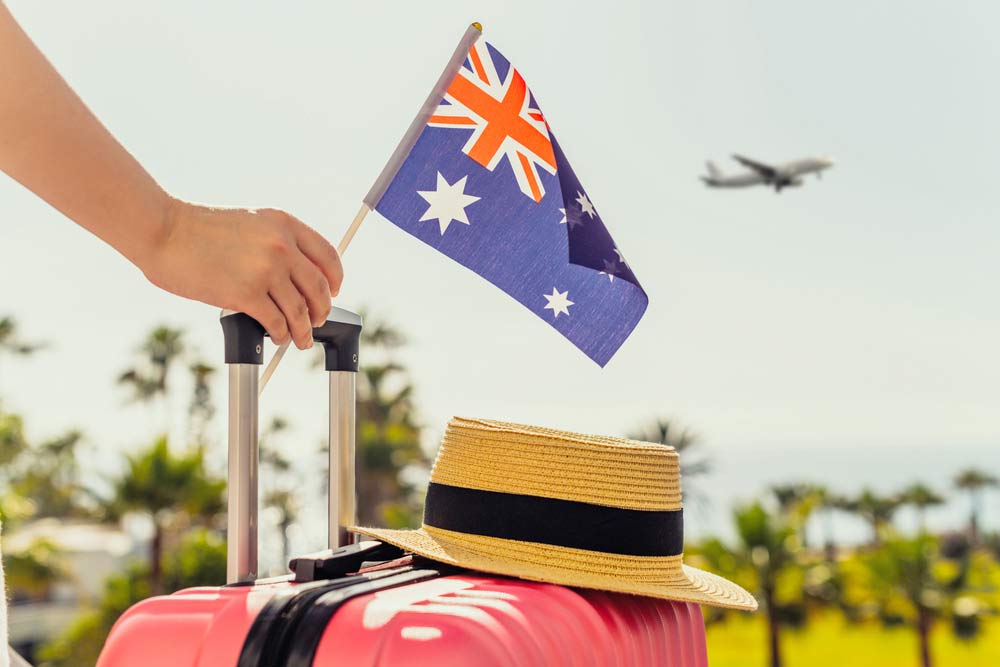 オーストラリア行きの旅行をイメージし、空港でスーツケースにオーストラリア国旗を掲げる手と、到着を待つ帽子の様子。