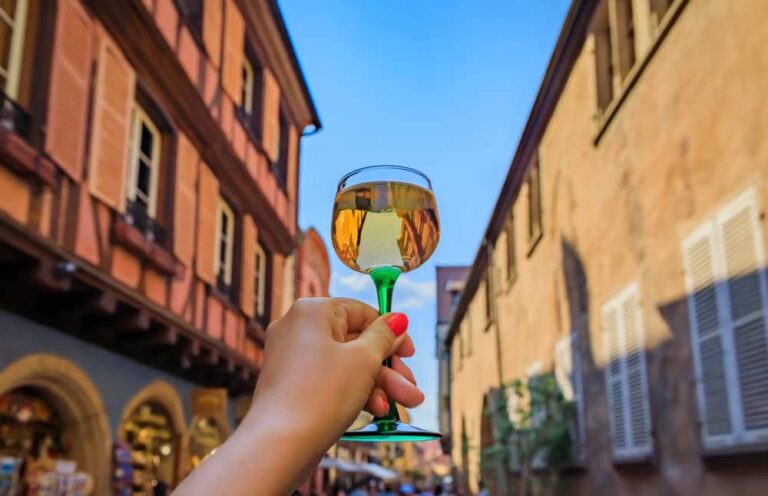 フランス・アルザスの街並みを背景に、緑のステム付きグラスで白ワインを掲げる手