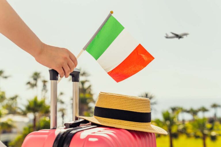 イタリアの国旗を持つ手と赤いスーツケース、ストローハットの上を飛行機が飛んでいる様子。旅行の出発を連想させるシーン。