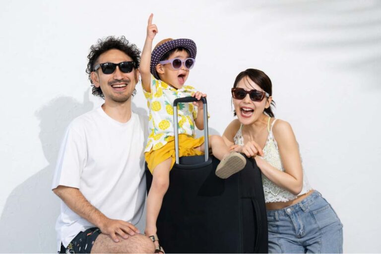 サングラスをかけた日本人家族がスーツケースとともに笑顔でポーズをとっている様子。夏の旅行を楽しむ雰囲気。