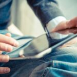 膝の上でタブレット端末を持ち、指で画面を操作する人の手元のクローズアップ。