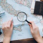 イタリアの地図を見ながら旅の計画を立てる手元。虫眼鏡、パスポート、コンパス、コーヒー、ユーロ紙幣が並ぶテーブル。
