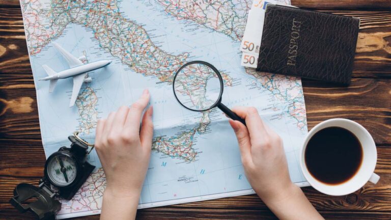 イタリアの地図を見ながら旅の計画を立てる手元。虫眼鏡、パスポート、コンパス、コーヒー、ユーロ紙幣が並ぶテーブル。
