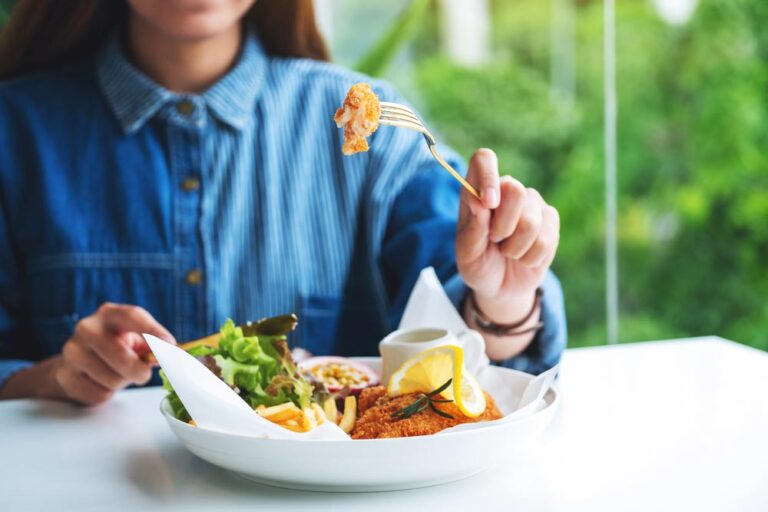 デニムシャツの女性が、サラダとフライドフィッシュのランチをフォークで食べている。