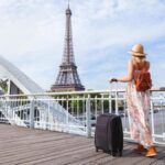 スーツケースを持ちセーヌ川沿いの橋からエッフェル塔を眺める女性旅行者