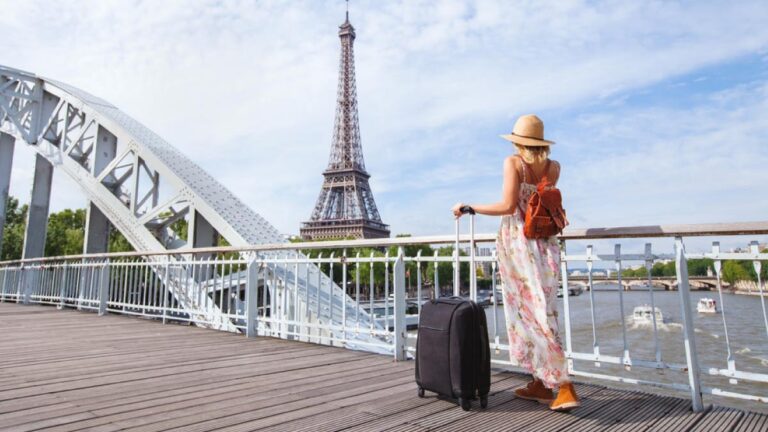 スーツケースを持ちセーヌ川沿いの橋からエッフェル塔を眺める女性旅行者