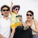 サングラスをかけた日本人家族がスーツケースとともに笑顔でポーズをとっている様子。夏の旅行を楽しむ雰囲気。