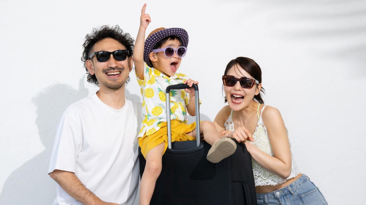 サングラスをかけた日本人家族がスーツケースとともに笑顔でポーズをとっている様子。夏の旅行を楽しむ雰囲気。