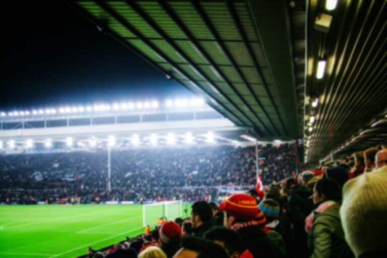 リバプールのサッカースタジアムで試合を観戦する観客たち。満員のスタンドと夜のライトに照らされて明るい緑のピッチ。