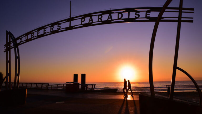 オーストラリア・ゴールドコーストの名所「サーファーズパラダイス」のゲート越しに眺める夕日。旅行者が散歩を楽しむ姿が シルエットで映し出され、現地ならではの特別な体験を感じられる景色。
