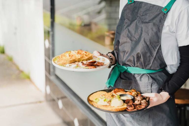 オーストラリアのカフェで提供されるブランチプレートを持つスタッフ。パンケーキや卵料理、ソーセージなどが並び、豊かなブランチ文化を感じられるシーン。