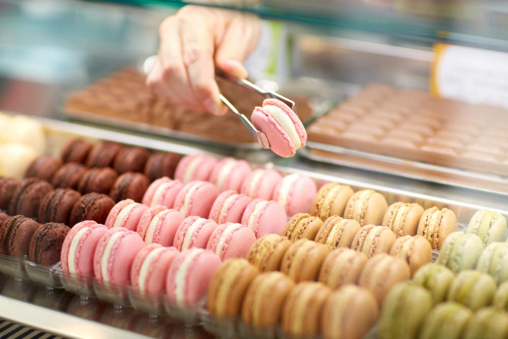 パリの洋菓子店で販売されているカラフルなマカロンをトングで選ぶ様子。ヨーロッパ土産に人気のスイーツの一例。