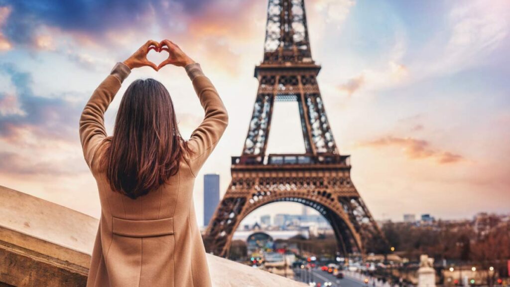 エッフェル塔を背景に、夕暮れの空に向かってハートの形を作る女性の後ろ姿。パリのロマンチックな雰囲気を感じる一枚。